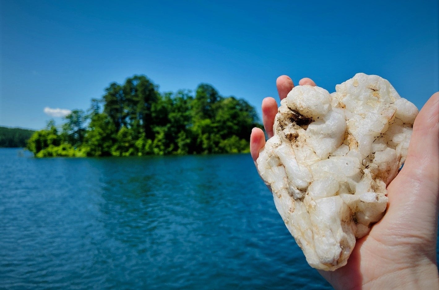 A person’s hand holds a large white crystal over a lake. An island sits in the background. 